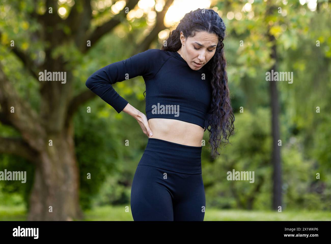 Eine hispanische Frau, die beim Sport in einem grünen Park Seitenschmerzen hat. Ihr Ausdruck zeigt Unwohlsein, während sie ihre Fitness-Routine unterbricht. Stockfoto