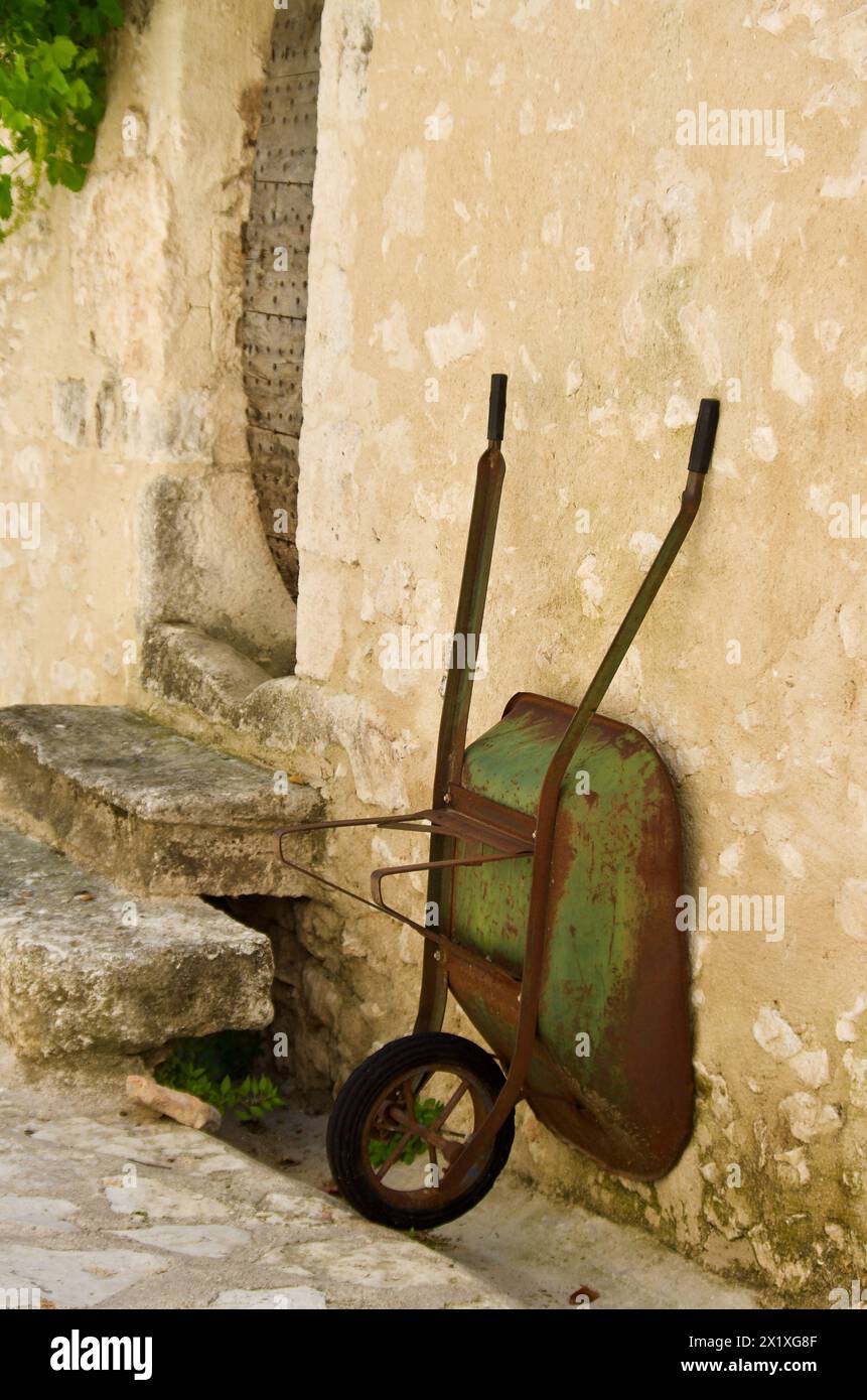 Alte, rostige Schubkarre steht vor einem Steingebäude vor einer Treppe in Frankreich. Stockfoto