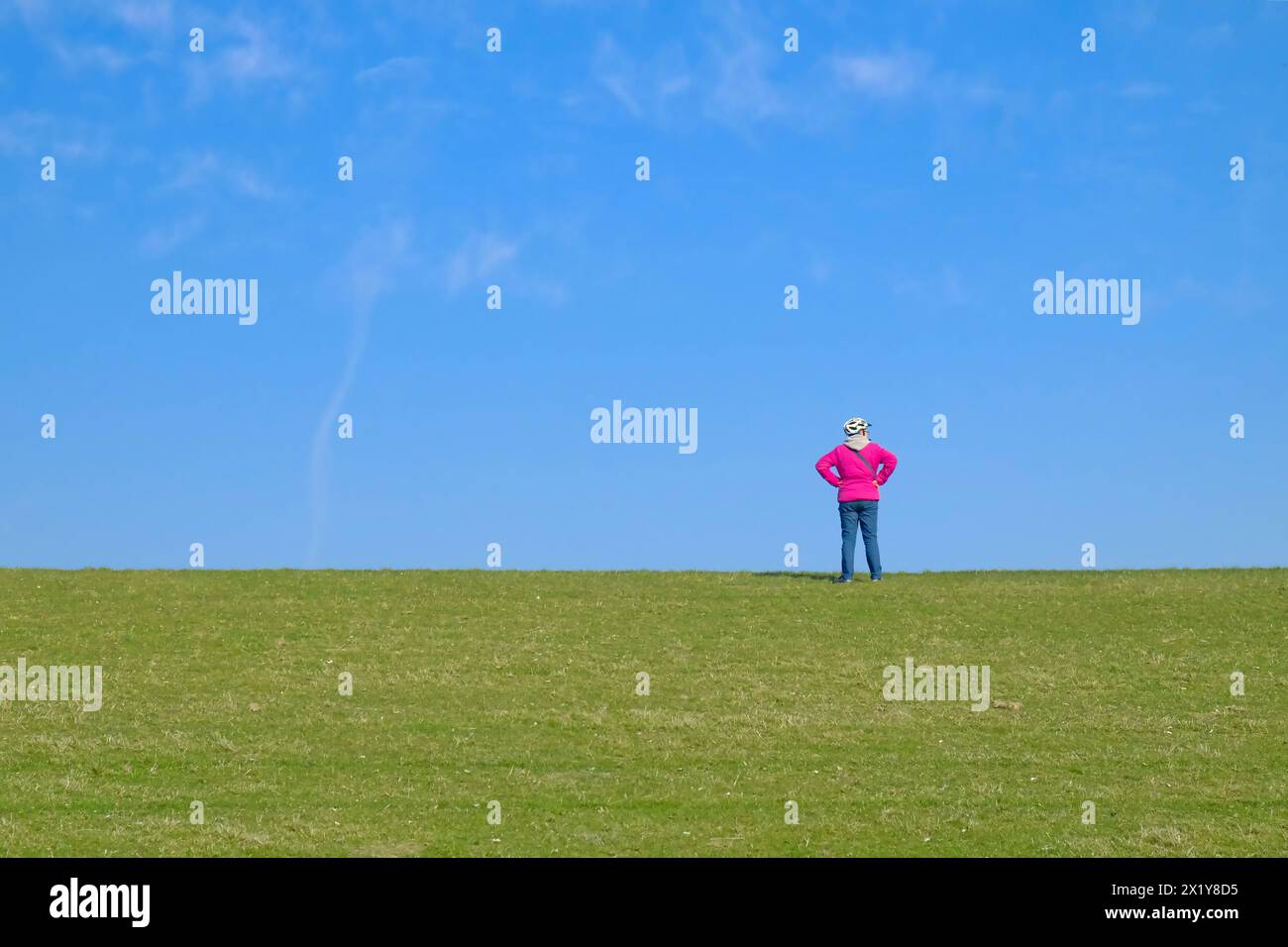 Radfahrer mit Helm und Pink Windbreaker allein auf Deich in Ostfriesland unter dem hellen blauen Himmel. Grün, rosa, Fahrradhelm, Ausblick, Deich, Sonne Stockfoto