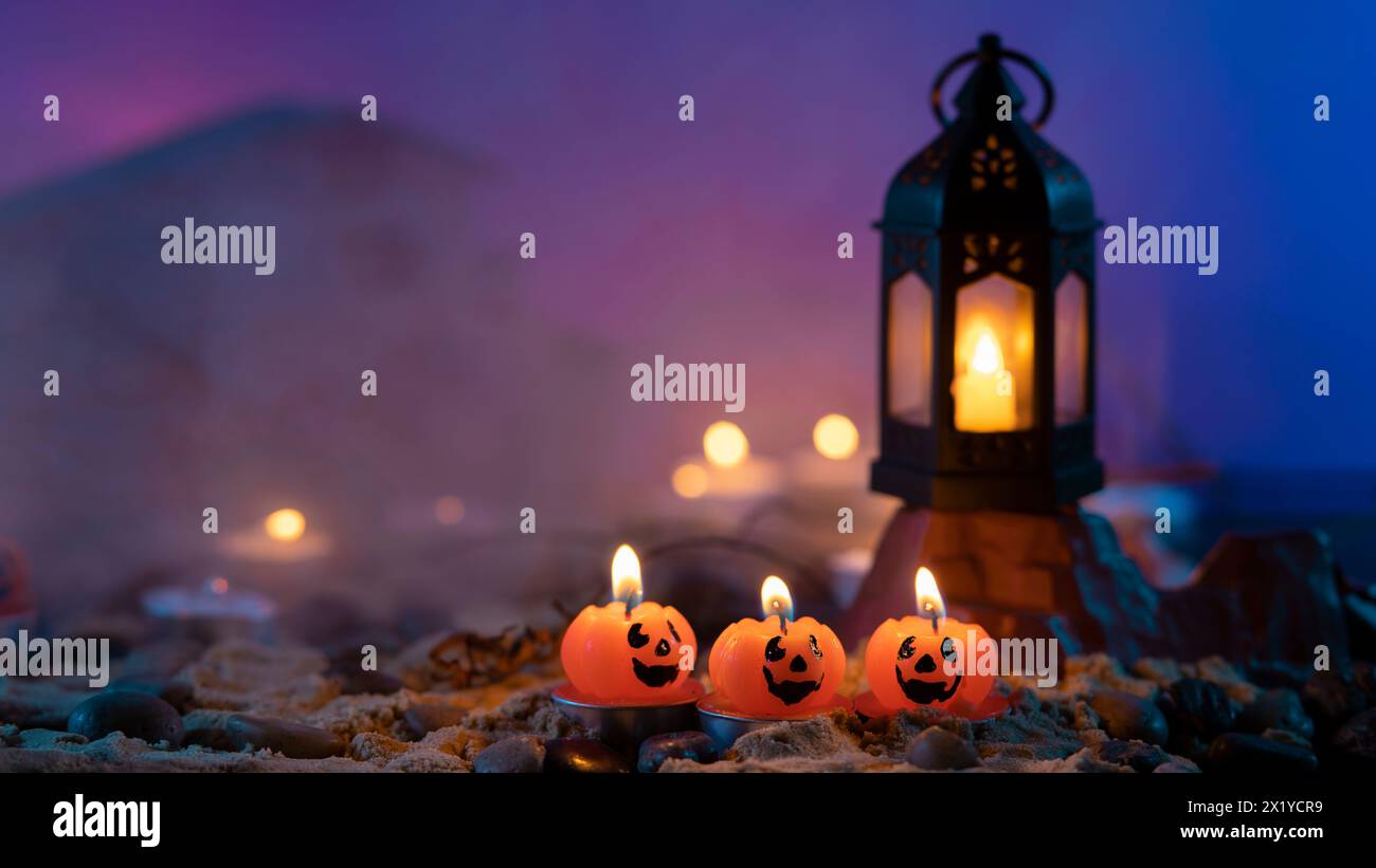 Halloween-Hintergrund, 3 Kerzen in Kürbisform im Vordergrund, eine alte Lampe auf einem Felsen vor einem blauen, unscharfen Hintergrund mit gelben Lichtern und Stockfoto