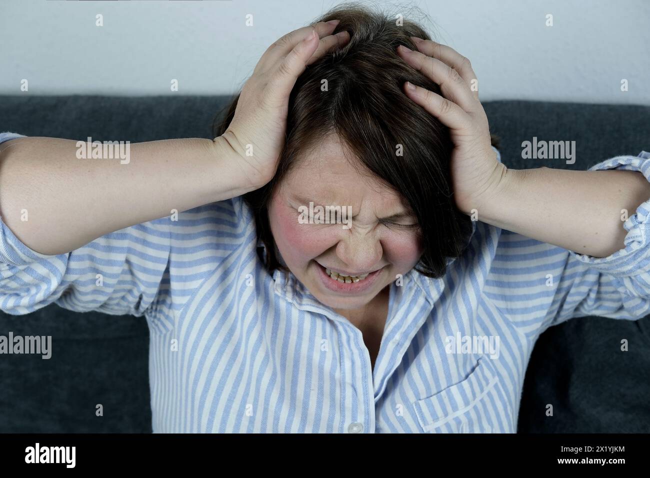 Erwachsene Frau sitzt auf einem Sofa, bedeckt mit einer Decke, klammert ihren Kopf in die Hände, Grimasse des Leidens im Gesicht, das Konzept des Migräneanfalls, p Stockfoto