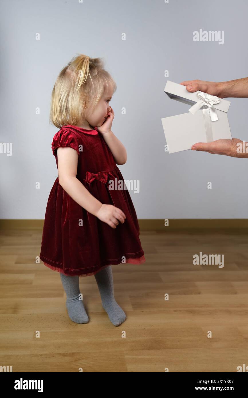 Das kleine blonde Mädchen 2 Jahre alt in einem roten Samtkleid steht im Profil im Raum, die Hände des Mannes zeigen ein Geschenk, das Konzept der Unschuld, Kindheit Stockfoto