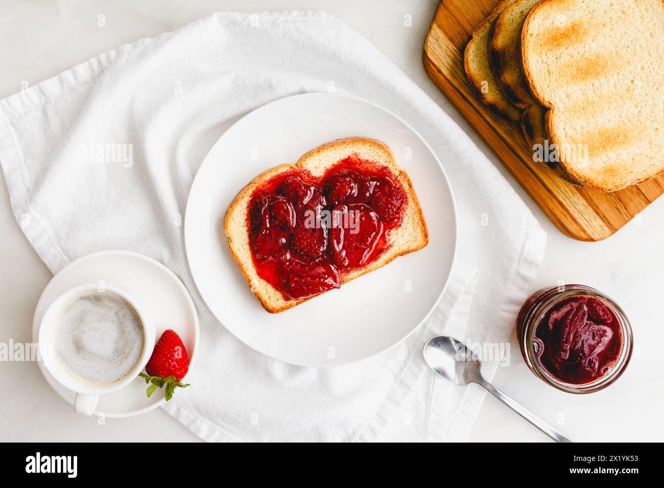 Frühstück mit Toast, Erdbeerkonserven und Cappuccino: Geröstetes Weißbrot und hausgemachte Beerenkonfitüren mit Kaffee auf weißem Tisch Stockfoto