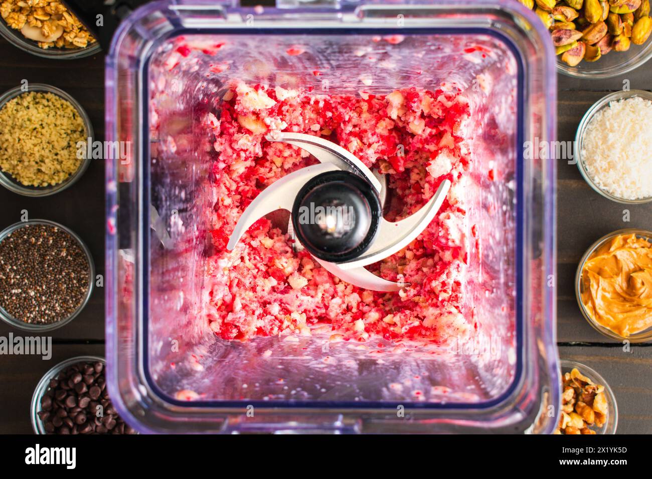 Gehackte gefrorene Erdbeeren und Bananen in einem Mixer: Direkt über dem mit gefrorenen Früchten gefüllten Pitcher, umgeben von Smoothie-Toppings Stockfoto