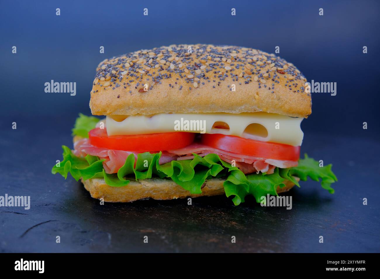 Nahaufnahme eines appetitlichen Sandwiches mit Reifen Tomatenscheiben, grünem Salat, Käse, Schinken, rotem Mohnbrötchen, schnellem Snack-Konzept, Fastfood-Zubereitung Stockfoto