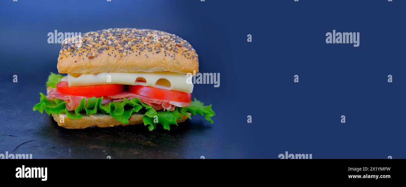 Nahaufnahme eines appetitlichen Sandwiches mit Reifen Tomatenscheiben, grünem Salat, Käse, Schinken, rotem Mohnbrötchen, schnellem Snack-Konzept, Fastfood-Zubereitung Stockfoto