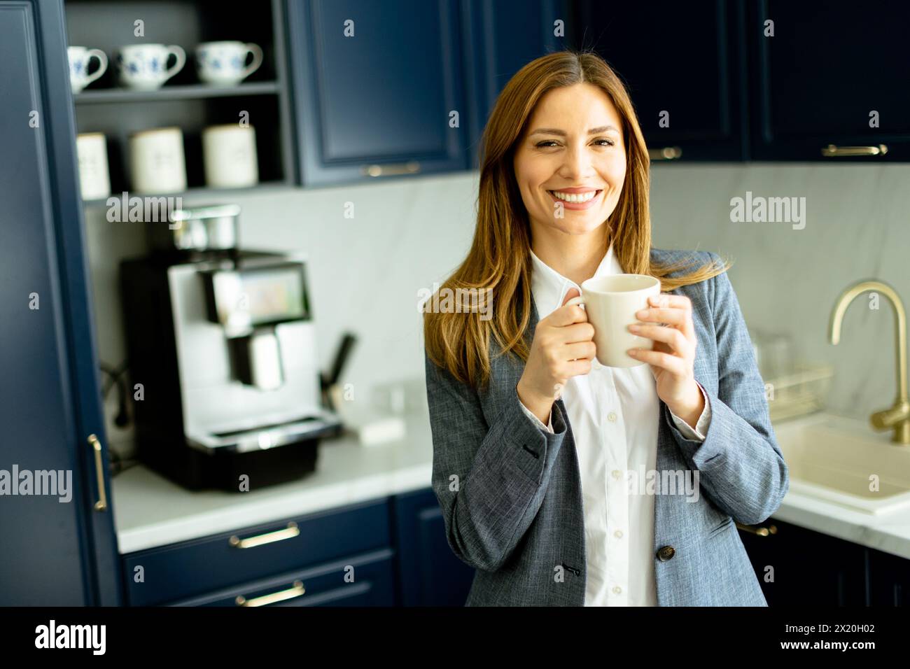 Eine lächelnde Frau in Geschäftskleidung, die eine Kaffeetasse hält und in einer stilvollen blauen Küche eine entspannende Pause einlegt. Stockfoto