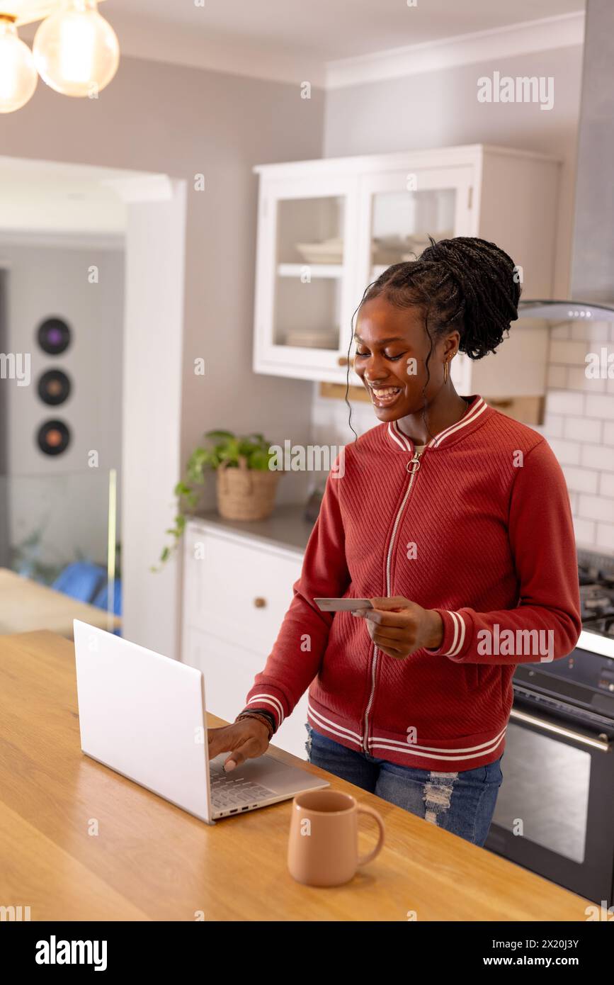 Afroamerikanische junge Frau, die Kreditkarten hält und in der Küche zu Hause einen Laptop benutzt Stockfoto