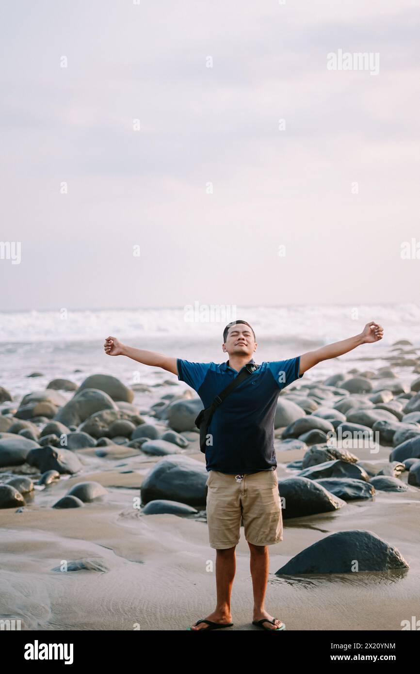 Ein zufriedener Mann in blauem T-Shirt und Shorts steht am Ufer, sein Blick auf den Horizont gerichtet und strahlt vor dem Hintergrund des beac Glück aus Stockfoto