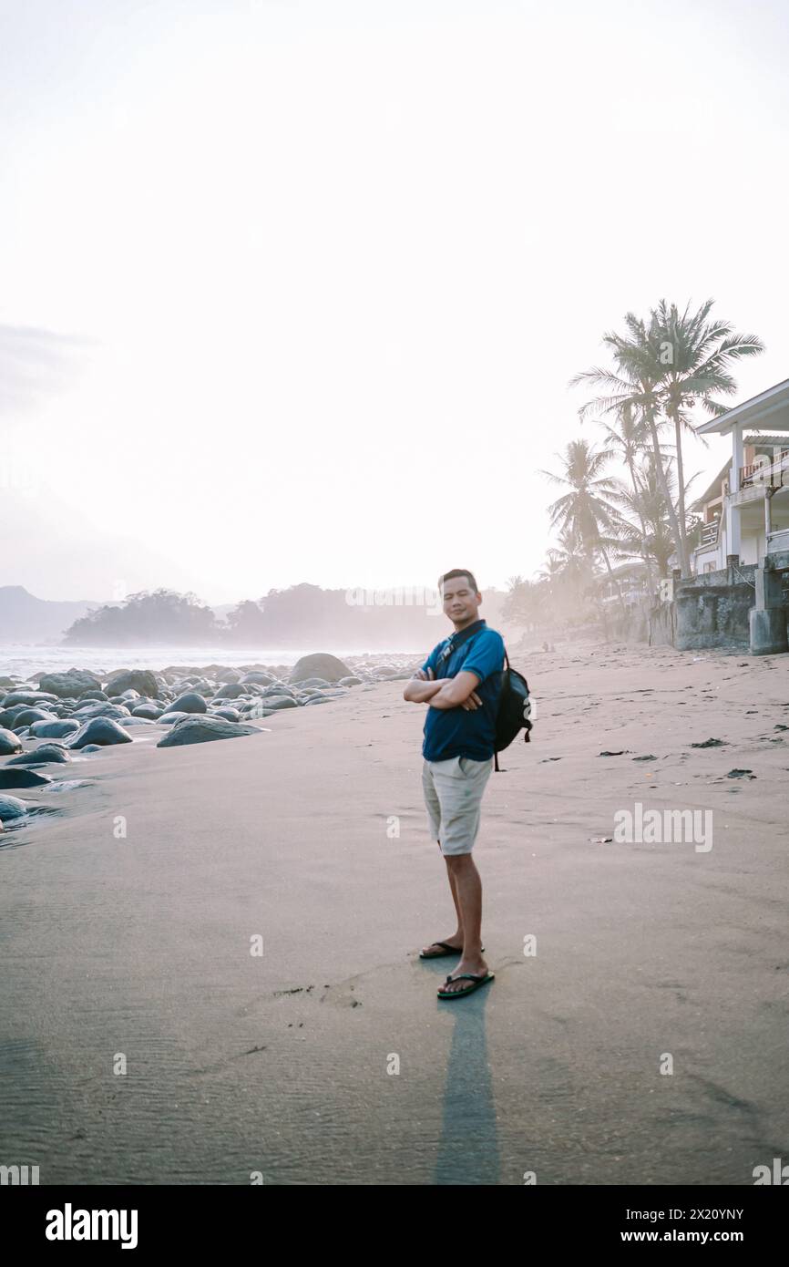 Ein zufriedener Mann in blauem T-Shirt und Shorts steht am Ufer, sein Blick auf den Horizont gerichtet und strahlt vor dem Hintergrund des beac Glück aus Stockfoto