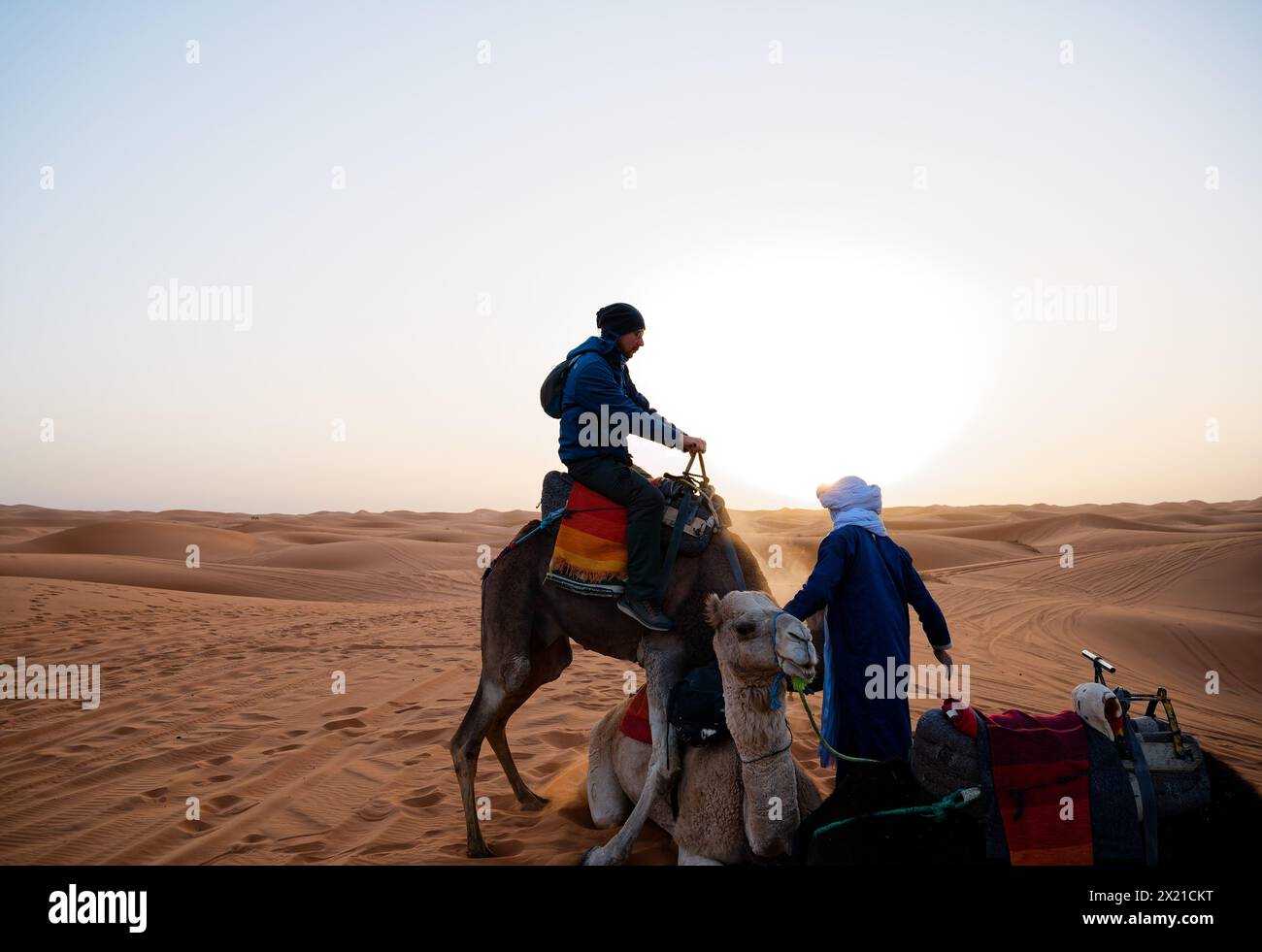 Ein Tourist mittleren Alters sitzt auf einem Dromedar und versucht, ein weiteres ruhendes Kamel zu besteigen, während ein ruhiger Handler gegen einen blendenden Sonnenaufgang eingreift. Stockfoto
