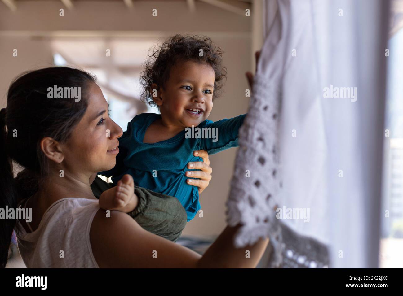 Die glückliche lateinamerikanische Mutter (30) und ihr Sohn (2) werden in den Armen gehoben und schauen beim Spielen zu Hause aus dem Fenster. Muttertagskonzept, alleinerziehende Mutter. Stockfoto