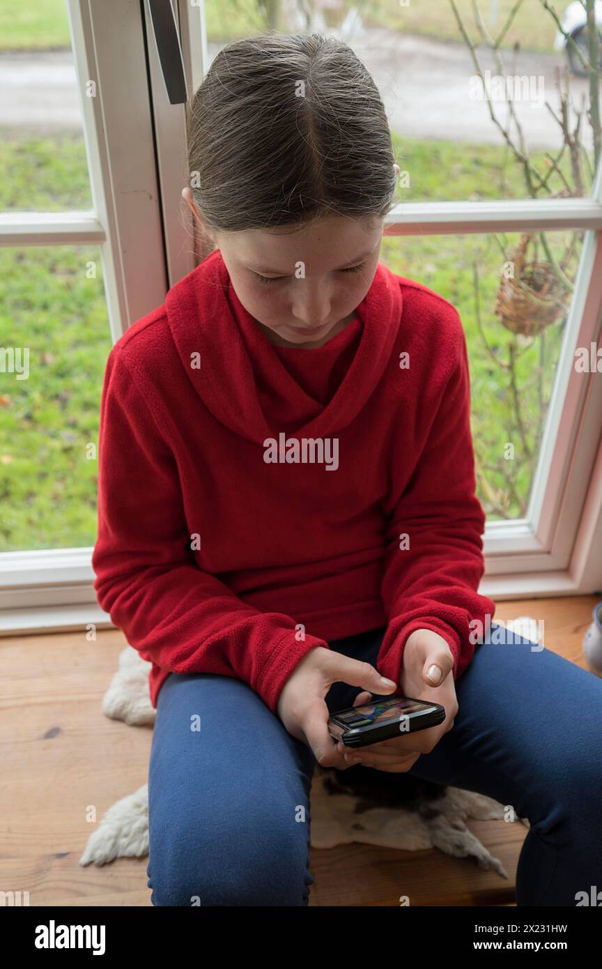 Mädchen, 10 Jahre alt, sitzt auf der Fensterbank mit Handy, Mecklenburg-Vorpommern, Deutschland Stockfoto