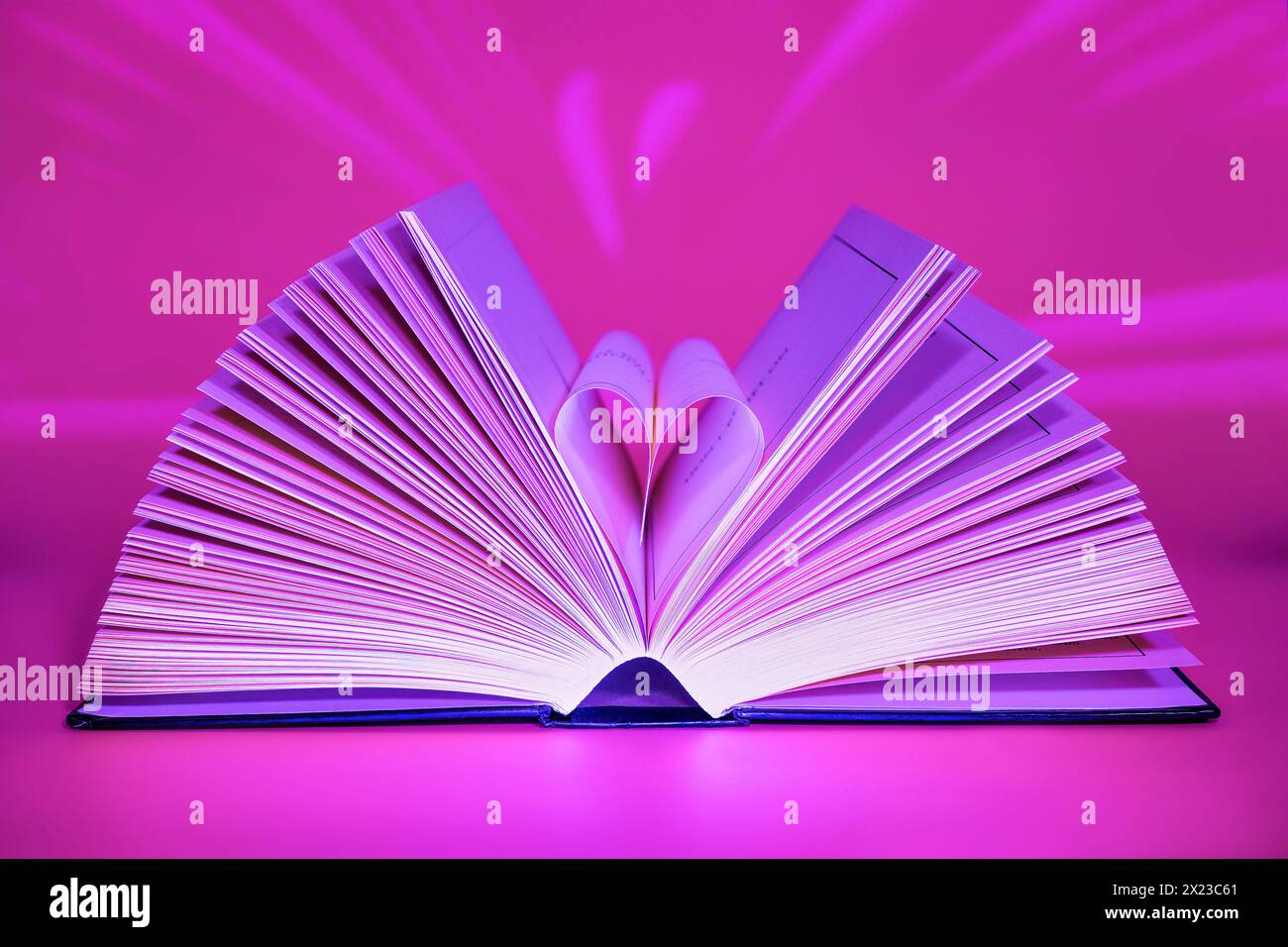 Offenes Buch auf rosafarbenem Hintergrund, beleuchtet von vorne mit interessanten Lichtmustern dahinter und Seiten in Herzform Stockfoto