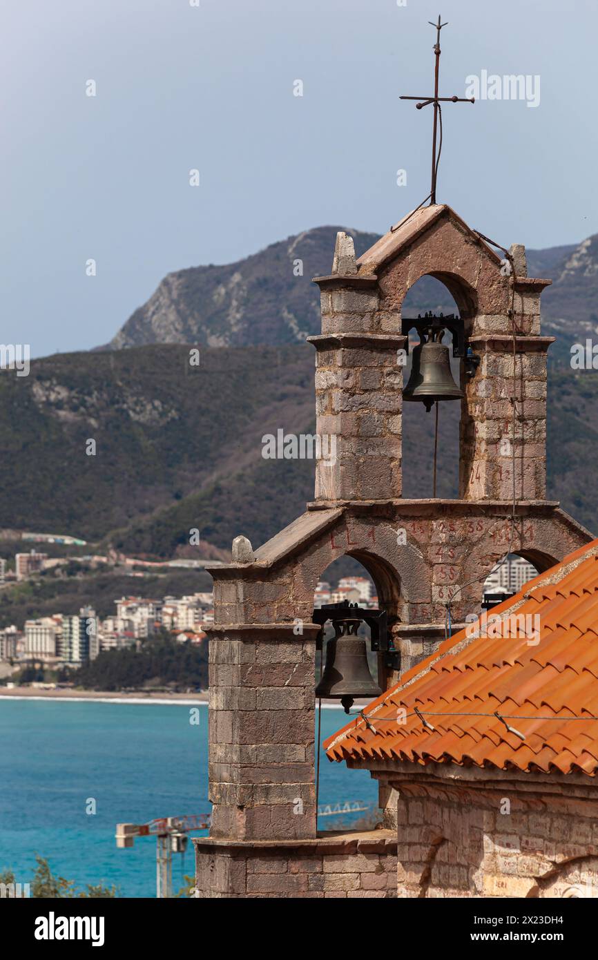 Glocken auf einem Steinkloster mit einem orangefarbenen Dach vor dem Hintergrund des Meeres und der Berge. Kloster oberhalb von Sveti Stefan in Montenegro. Egorova-Pfad. Stockfoto