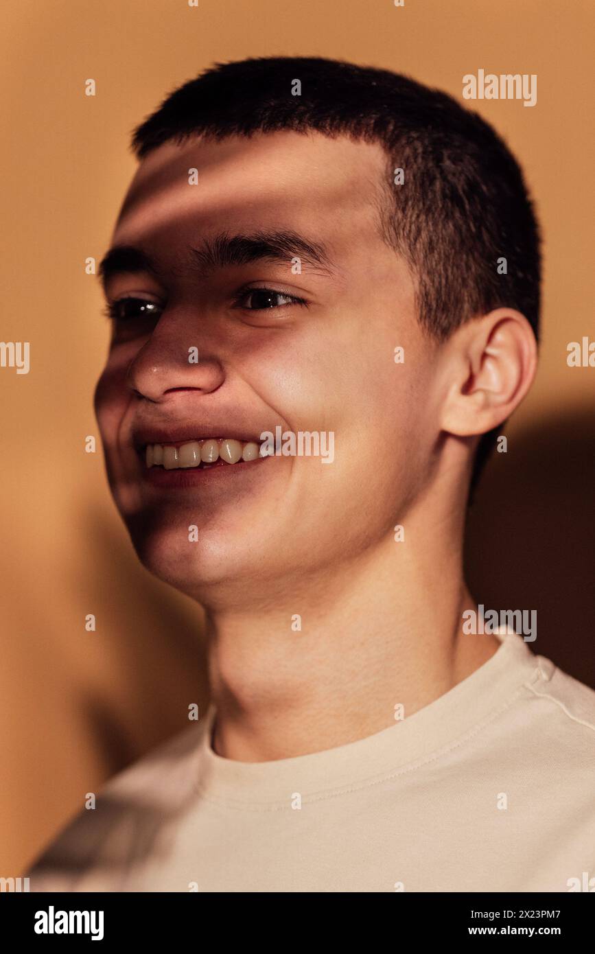 Nahporträt eines männlichen nachdenklichen Teenagers im warmen Innenlicht. Ein nachdenklicher junger Mann im Profil mit Schatten eines Blattes, das auf seinem Gesicht spielt. Beige Stockfoto