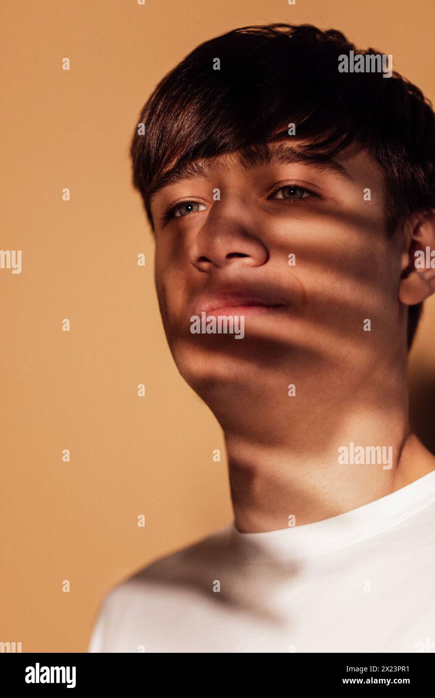 Nahporträt eines männlichen nachdenklichen Teenagers im warmen Innenlicht. Ein nachdenklicher junger Mann im Profil mit Schatten eines Blattes, das auf seinem Gesicht spielt. Beige Stockfoto