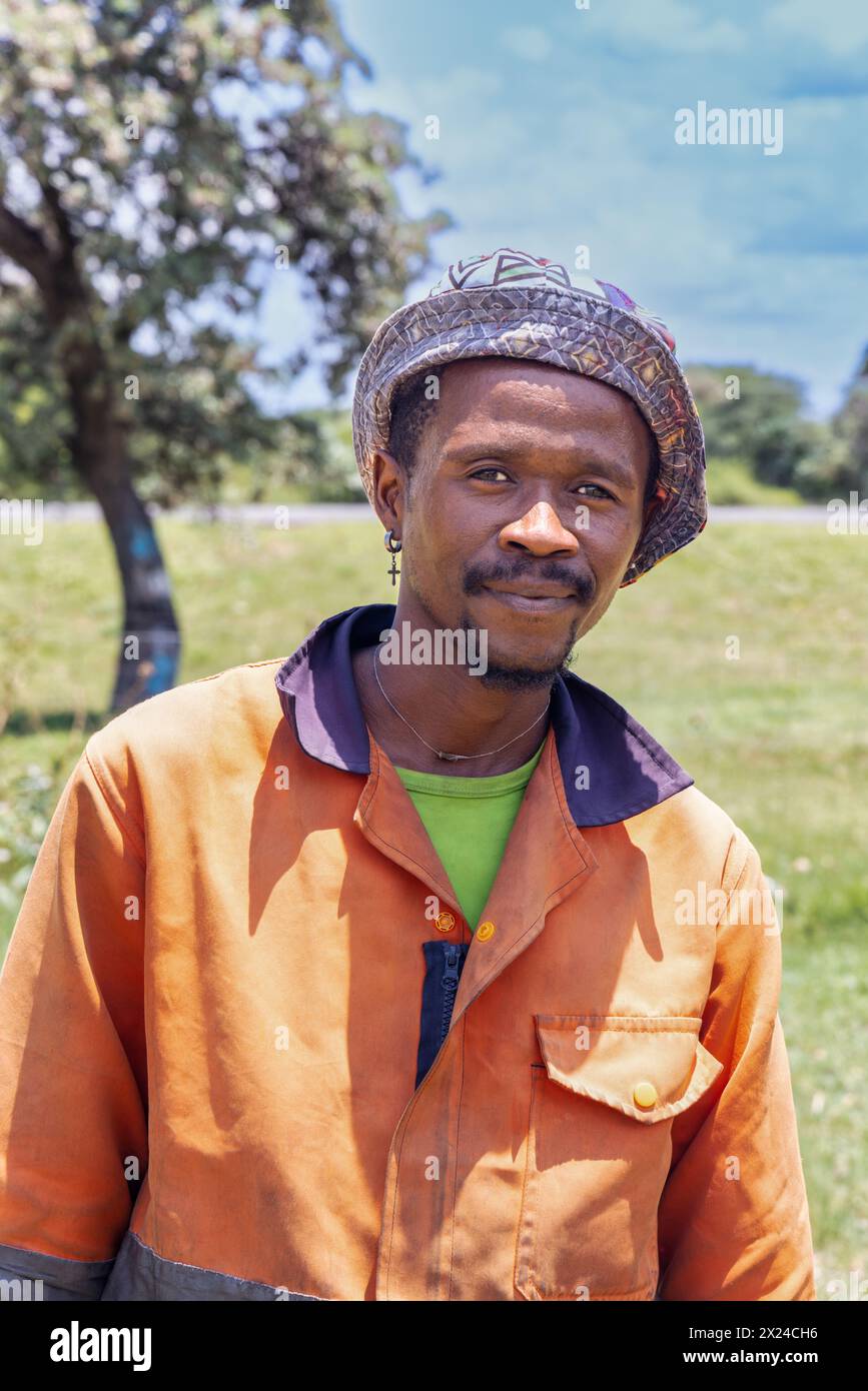 Junger afrikanischer Mann im Dorf, trägt orange Arbeitskleidung und als Beanie, Green Field und Baum Stockfoto
