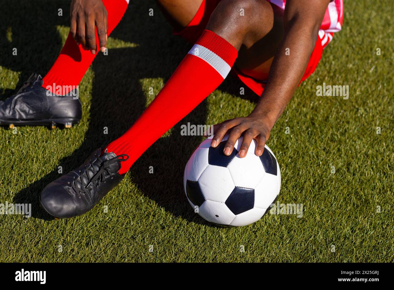 Afroamerikanischer junger männlicher Athlet, der draußen auf dem Rasen sitzt und einen Fußball hält Stockfoto