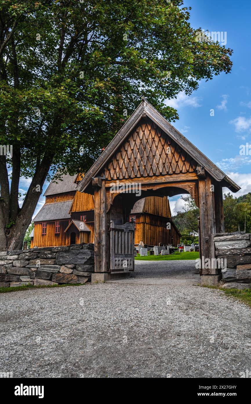 Ein hölzernes Tor führt zur historischen mittelalterlichen Ringebu Stave Kirche, umgeben von Grabsteinen vor einem sommerblauen Himmel mit Wolken Innlandet, Norwegen Stockfoto