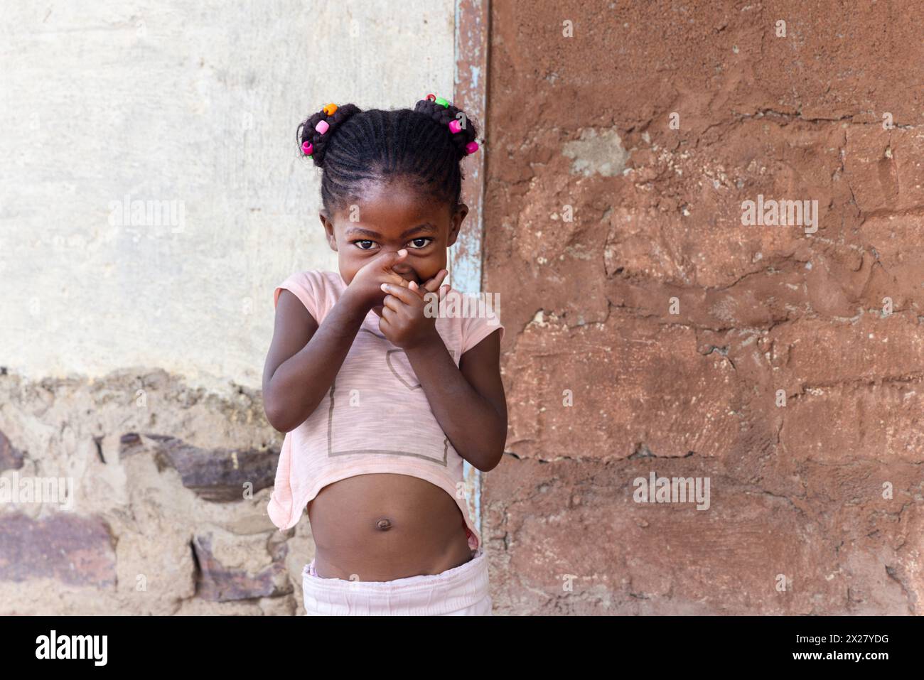 afrikanisches Mädchen mit Zöpfen und Perlen, das lustige Gesichter vor einer Wand in der Township zieht Stockfoto