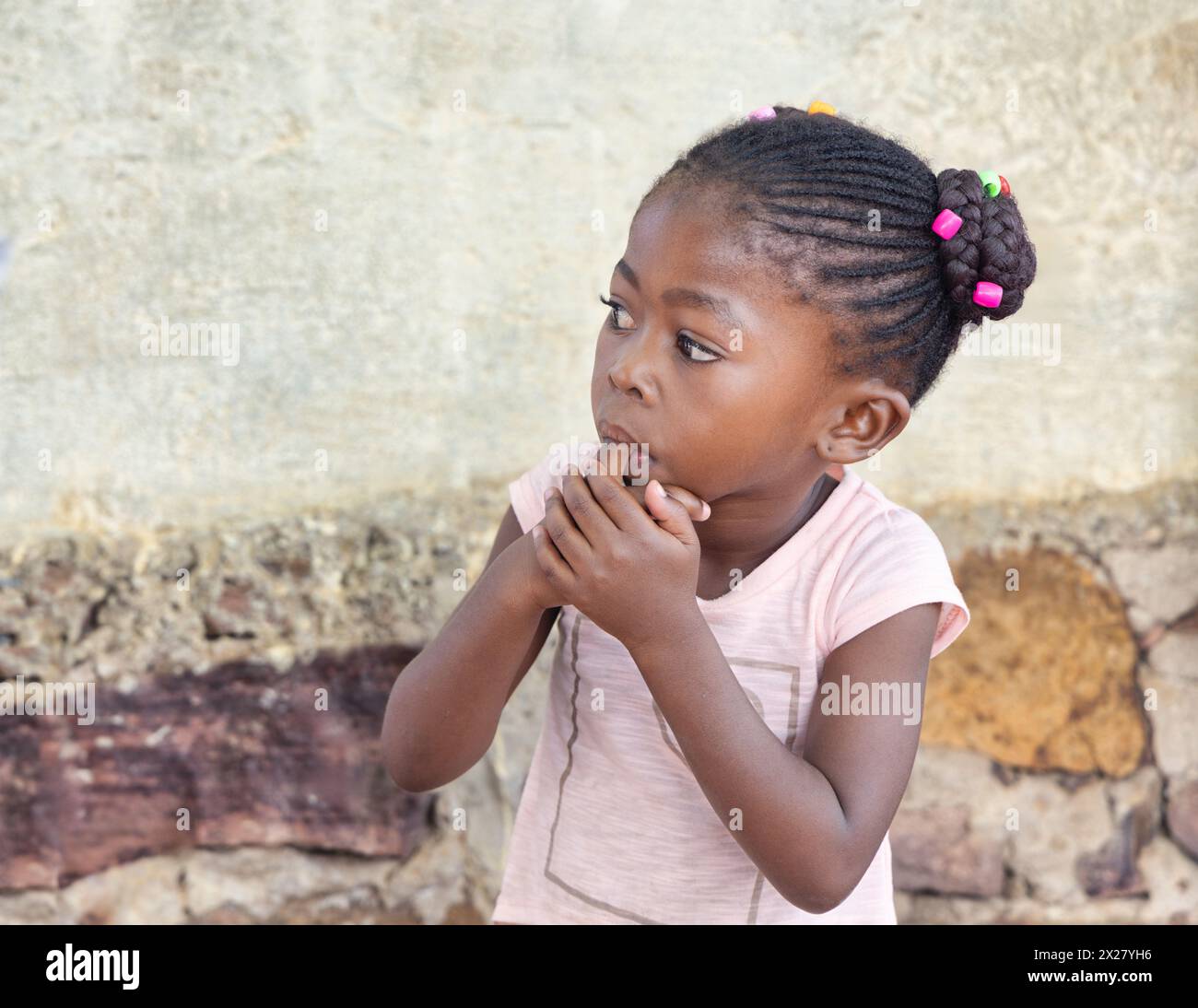 afrikanisches Mädchen mit Zöpfen und Perlen, die ihren Finger im Mund halten, vor einem Haus in der Township Stockfoto