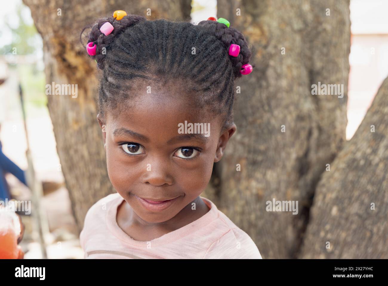 afrikanisches Mädchen mit Zöpfen und Perlen, vor einem Baum in der Gemeinde Stockfoto