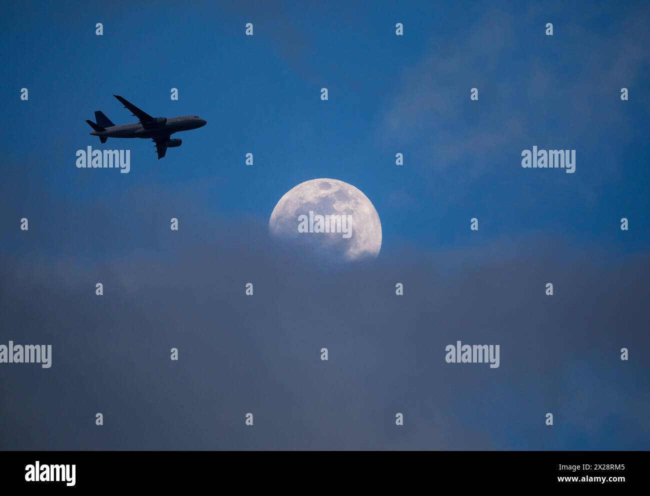 Ein kommerzielles Flugzeug, das sich auf einem Flughafen befindet, überquert einen Vollmond, der in der Abenddämmerung teilweise von niedrigen Wolken verdeckt ist. Stockfoto