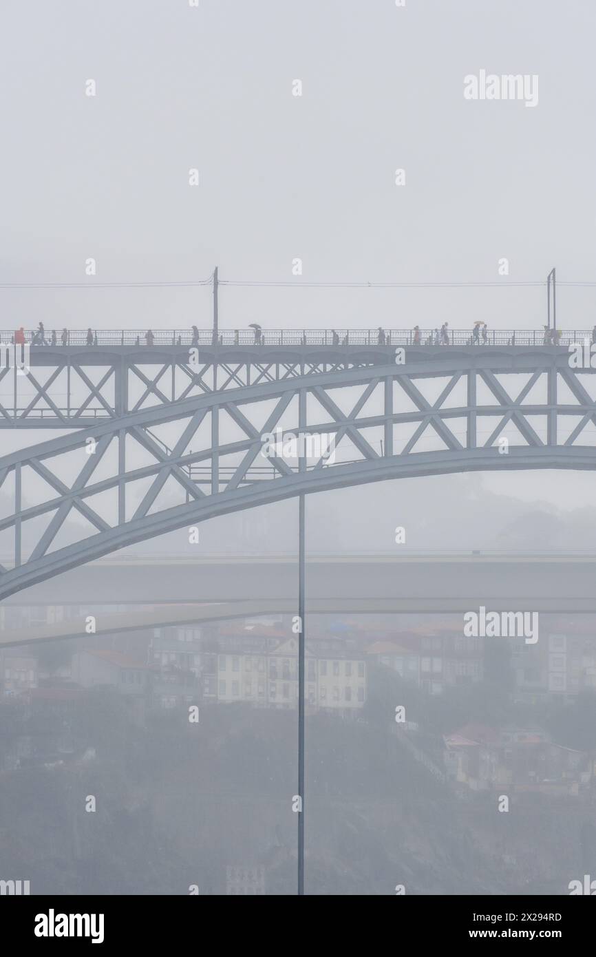Menschen mit Regenschirmen und Regenmänteln gehen auf der oberen Plattform der Don Luis I Stahlbrücke und Autos fahren auf der unteren Plattform mit einer Person Stockfoto