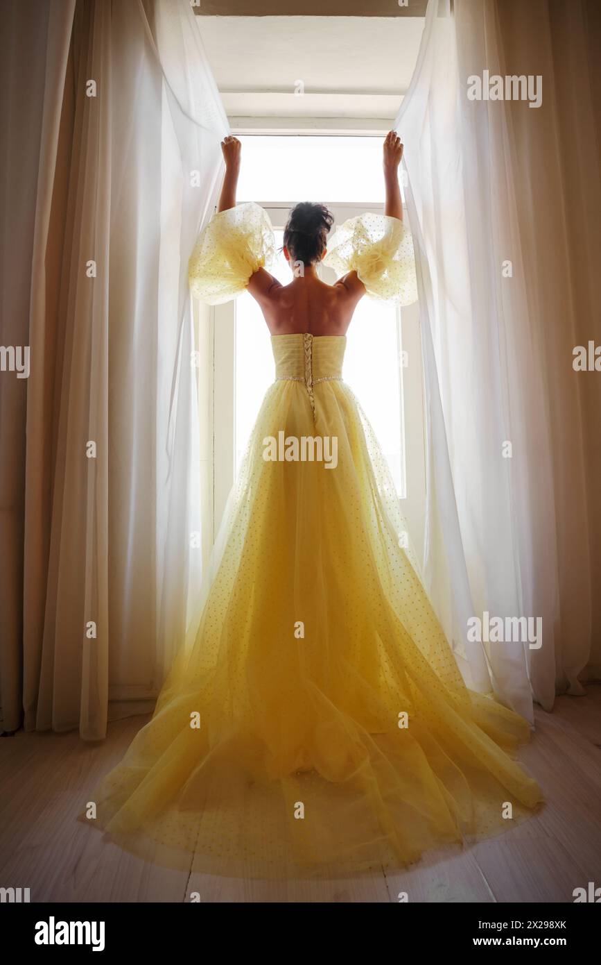 Die Silhouette einer Frau in einem goldenen, luxuriösen Kleid vor dem Hintergrund eines Fensters hält einen Vorhang mit den Händen. Elegante Dame in gelber Seide Stockfoto