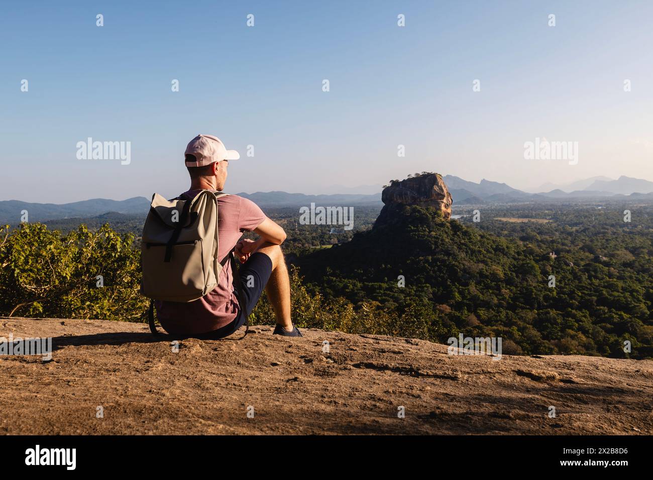 Mann mit Rucksack, der auf Felsen sitzt und die Landschaft betrachtet. Wunderschöne Landschaft mit Sigiriya-Felsen. Alleinreisender in Sri Lanka. Stockfoto
