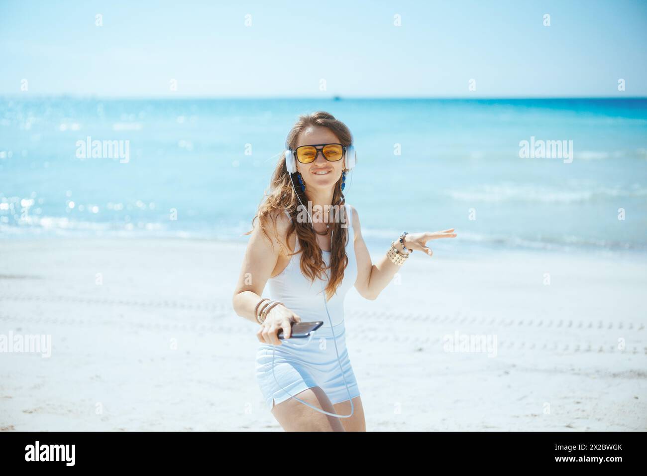 Lächelnde, elegante Frau an der Meeresküste mit Kopfhörern und tanzendem Smartphone. Stockfoto