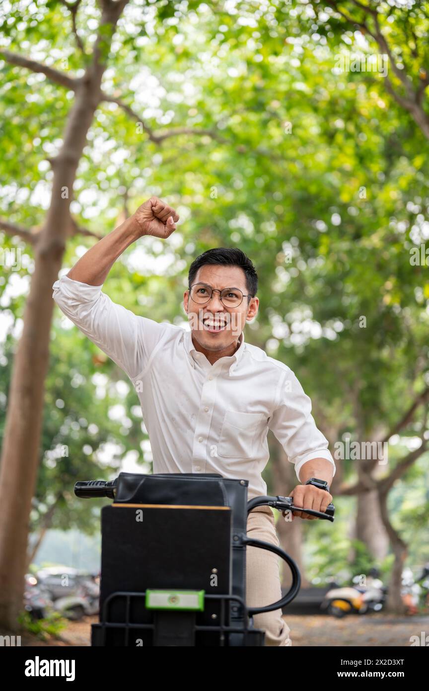Ein aufgeregter asiatischer Geschäftsmann fährt triumphierend auf dem Fahrrad, hebt seine Faust, hat ein Ziel erreicht oder gute Nachrichten erhalten. Stockfoto