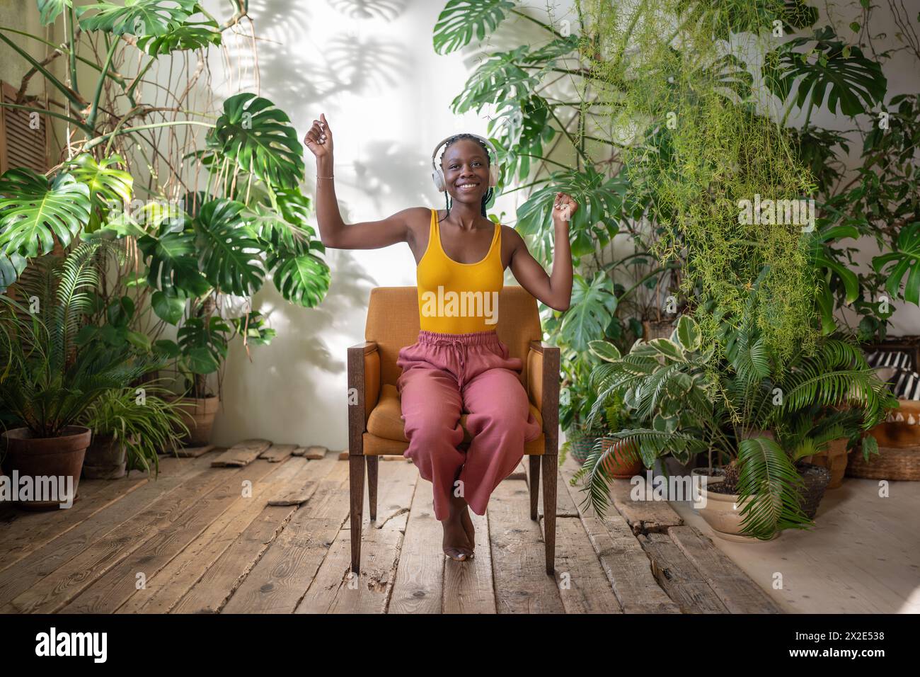 Fröhliches glückliches schwarzes Mädchen in Kopfhörern hört Musik zwischen tropischen Zimmerpflanzen lächelnd sitzt auf dem Stuhl. Stockfoto