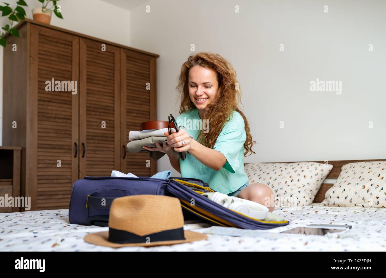 Wir freuen uns auf Reisen. Lockige Frau sitzt zu Hause auf ihrem Bett und packt ihre Reisetasche für den Sommerurlaub. Stockfoto