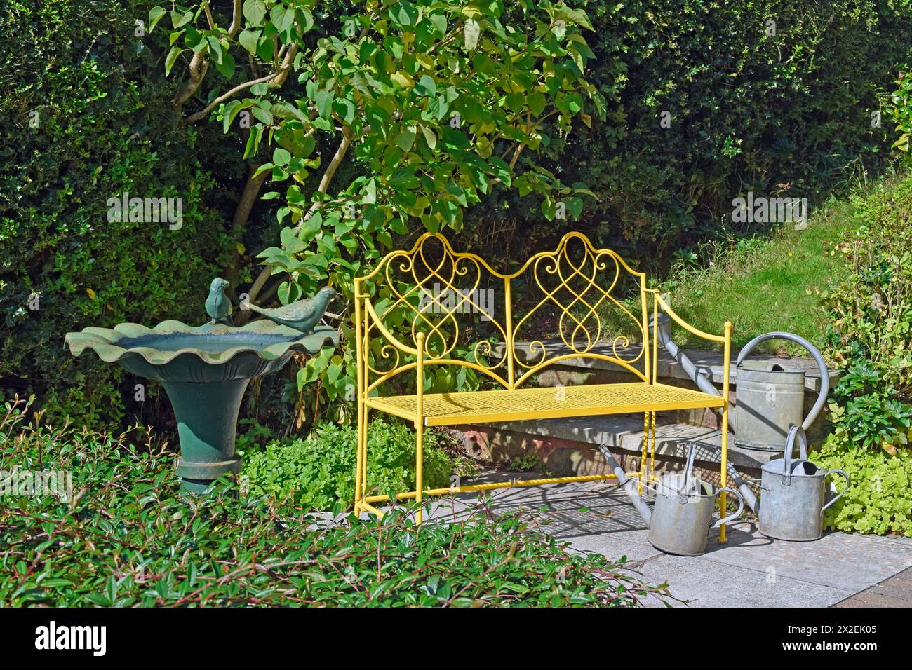 Botanik, Gelb verzierte Metallbank in einem Garten mit Metall-Vogelbad und Gießkannen, ZUSÄTZLICHE RECHTE-CLEARANCE-INFO-NICHT-VERFÜGBAR Stockfoto