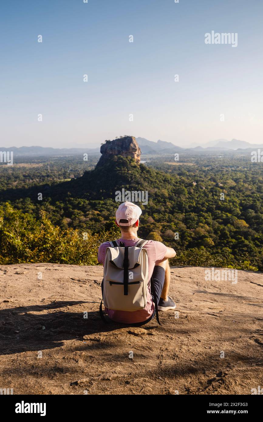 Mann mit Rucksack, der auf Felsen sitzt und die Landschaft betrachtet. Wunderschöne Landschaft mit Sigiriya-Felsen. Alleinreisender in Sri Lanka. Stockfoto