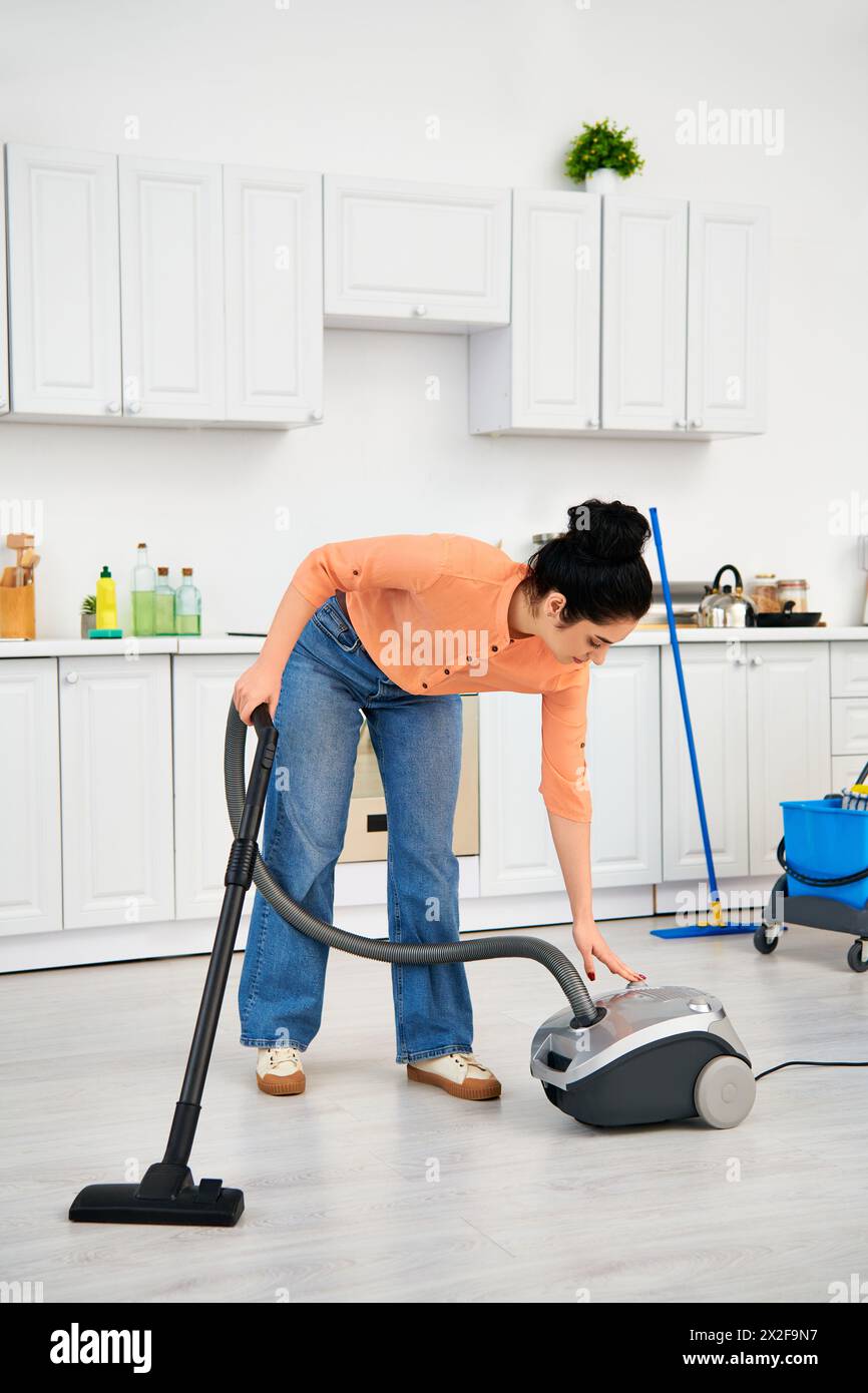 Eine stilvolle Frau in lässiger Kleidung reinigt leidenschaftlich ihren Küchenboden mit einem Staubsauger. Stockfoto