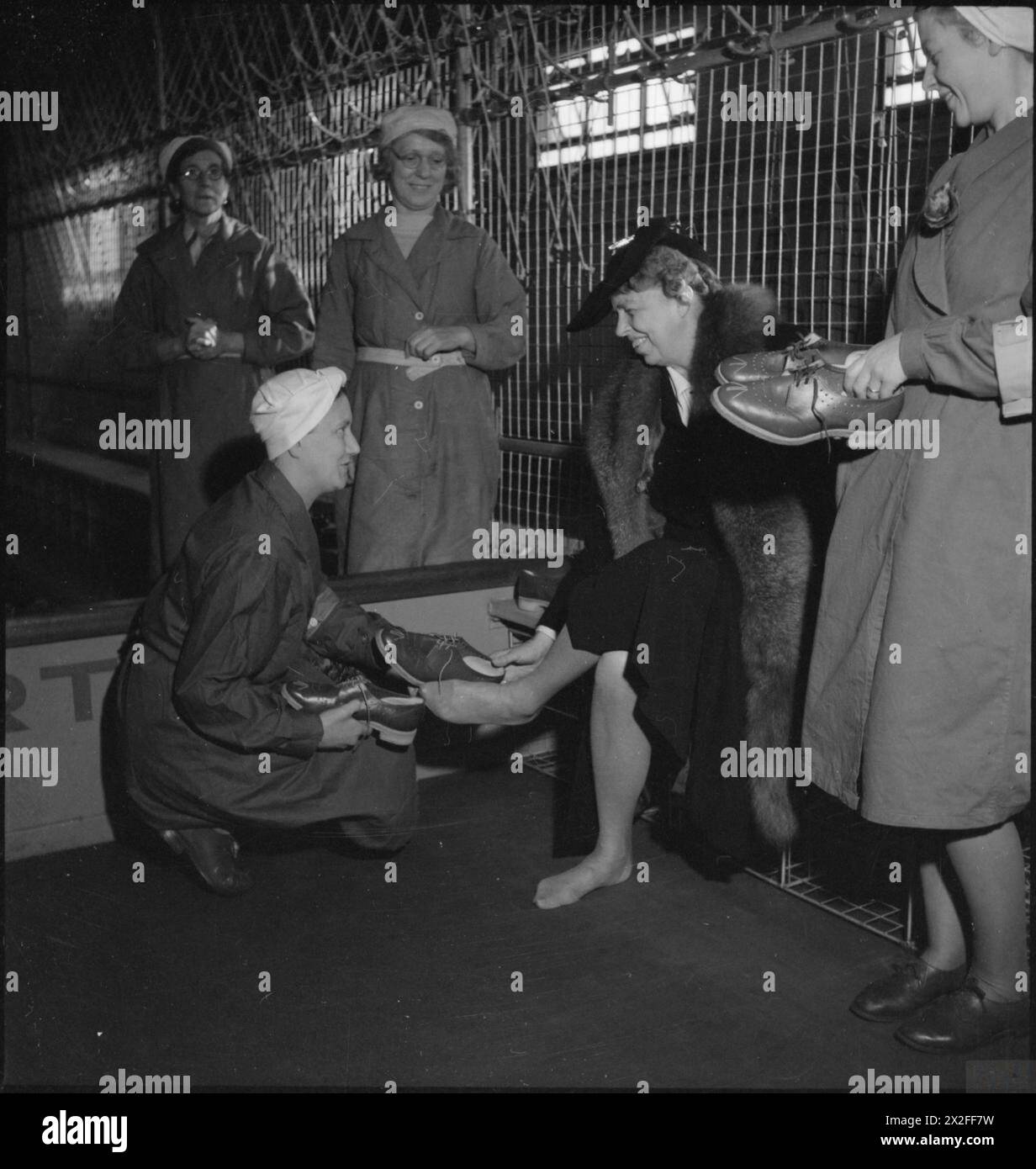 FRAU ROOSEVELT BESUCHT Eine KÖNIGLICHE KAMPFMITTELFABRIK in Großbritannien, 1942 - Eleanor Roosevelt wird von einem Munitionsarbeiter mit Spezialschuhen ausgestattet, bevor sie in den Gefahrenbereich einer britischen Royal Ordnance Factory gelangen kann. Der Bereich der Fabrik, in dem die Arbeiter von ihrer Außenkleidung (die „schmutzige“ Seite) in ihre Fabrikkleidung wechseln und über eine niedrige Barriere (deutlich sichtbar) auf die „saubere“, Fabrikseite traten, wird als „Umzugshaus“ bezeichnet. Auf der „sauberen“ Seite wurden strenge Vorschriften angewandt, da die Arbeiten bei der Abfüllung von Fabriken äußerst gefährlich waren Stockfoto