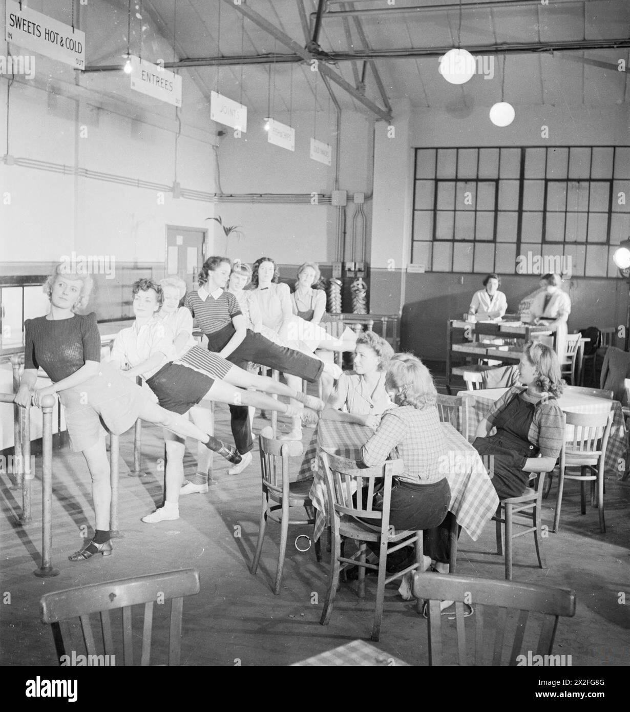 FACTORY BALLET: WARTIME FACTORY ENTERTAINMENT, 1942 – weibliche Fabrikarbeiter, darunter Florence Simpson, Winifred Bennett und ihre Schwester Dolly, üben am Ende ihrer Schicht einige Ballettzüge in der Kantine der Flugzeugfabrik, in der sie arbeiten, irgendwo in Großbritannien. Sie benutzen die Bars, die die Warteschlangen der Gäste trennen, als Barren. Über ihren Köpfen sind die Zeichen für heiße und kalte Süßigkeiten, Hauptgerichte und andere Lebensmittel deutlich zu erkennen. Rechts neben dem Foto sitzen mehrere andere Frauen und beobachten die Tänzerinnen, wie sie sich nach dem Essen entspannen Stockfoto