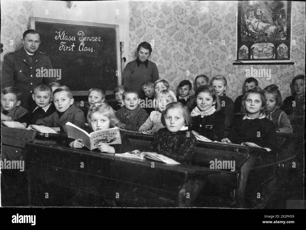 Polnische Kinder lernen 1945 in einem Hanauer Flüchtlingslager in Deutschland lesen, unter der Aufsicht von UNRRA-Mitarbeitern und einem Priester, der Auschwitz überlebte und ein polnisches Alphabet namens Elementarz hielt. Stockfoto