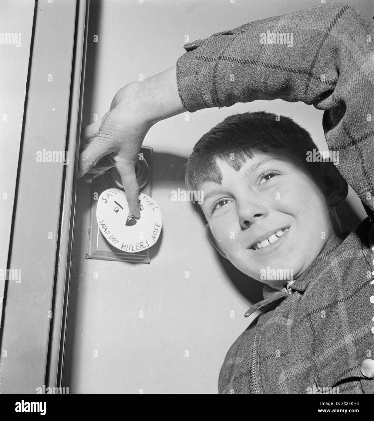 KRAFTSTOFFVERBRAUCH IN DER WOHNUNG, GROSSBRITANNIEN, C 1942 - Ein kleiner Junge spielt seine Rolle bei der Kraftstoffeinsparung, indem er einen Lichtschalter ausschaltet. Über dem Lichtschalter steht eine Zeichnung mit der Aufschrift „Kraftstoff sparen: Hitlers Nase abreißen“. Stockfoto