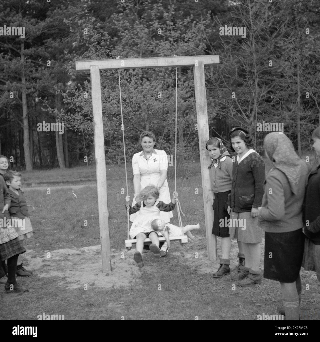 In Bergen-Belsen spielt eine russische Freiwillige Krankenschwester aus Lager Nr. 1 mit Kindern auf Schaukeln, die von den Royal Electrical and Mechanical Engineers (R.E.M.E) installiert wurden. Stockfoto