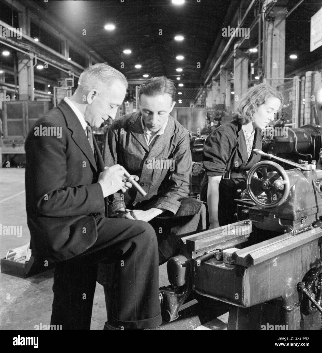 A DAY IN THE LIFE OF A SHOP Steward: FACTORY WORK IN BRITAIN, 1942 – Shop Steward Richard Sainsbury (Mitte) berät sich mit Henry Howard, dem Vorarbeiter des Werkzeughauses, über das Problem, das der Arbeiter Megan Beynon ihnen vorstellte. Megan hat wieder an ihrer Drehmaschine gearbeitet, während sie versuchen, das Problem zu lösen Stockfoto