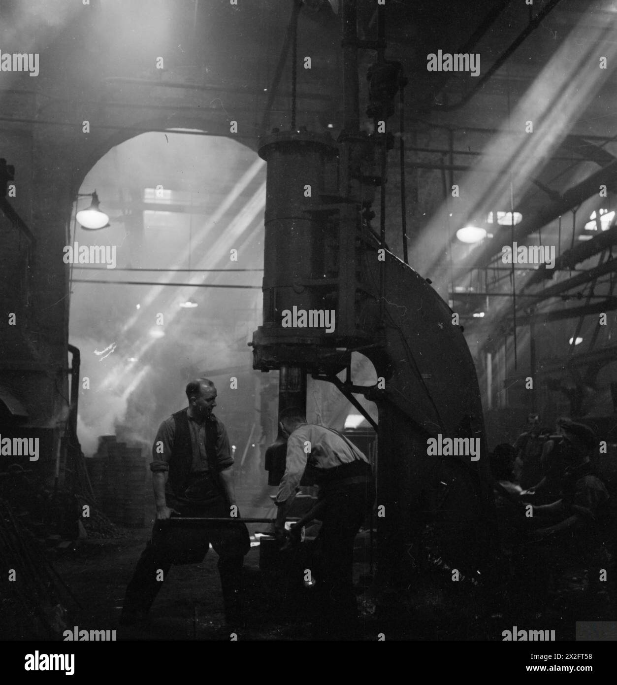 Zwei Schmiede arbeiten 1942 in einer britischen Schmiede und Formen Teile für Matilda-Panzer. Lichtwellen beleuchten die dunkle, rauchige Atmosphäre der Tankfabrik während der Kriegszeit. Stockfoto