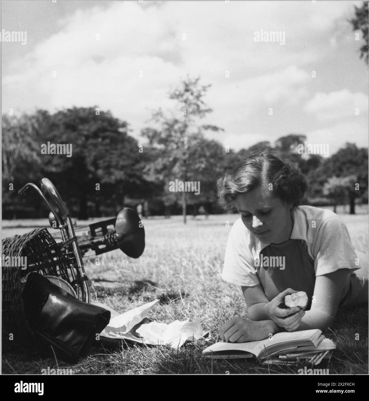 LONDON PARKS: UNTERHALTUNG UND ENTSPANNUNG IM HERZEN DER STADT, LONDON, ENGLAND, 1943 - Ein junger Kriegsarbeiter liest und isst einen Apfel in der Sonne im Hyde Park. Neben ihr ist ihr Fahrrad, mit dem sie sie von ihrem Kriegsjob zu einem Picknick in der Sonne transportiert hat. Laut der Originalüberschrift kommt sie im Sommer oft zum Mittagessen in den Park Stockfoto