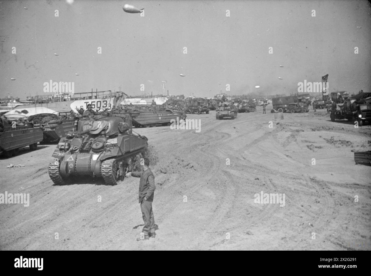 Panzer und Ausrüstung entladen an den Stränden der Normandie während der Operation Overlord, Juni 1944; Soldat leitet den Panzer; LCT 503 und LCT 7096 an Bord; DUKWs, LKWs und Sperrballons sichtbar. Stockfoto