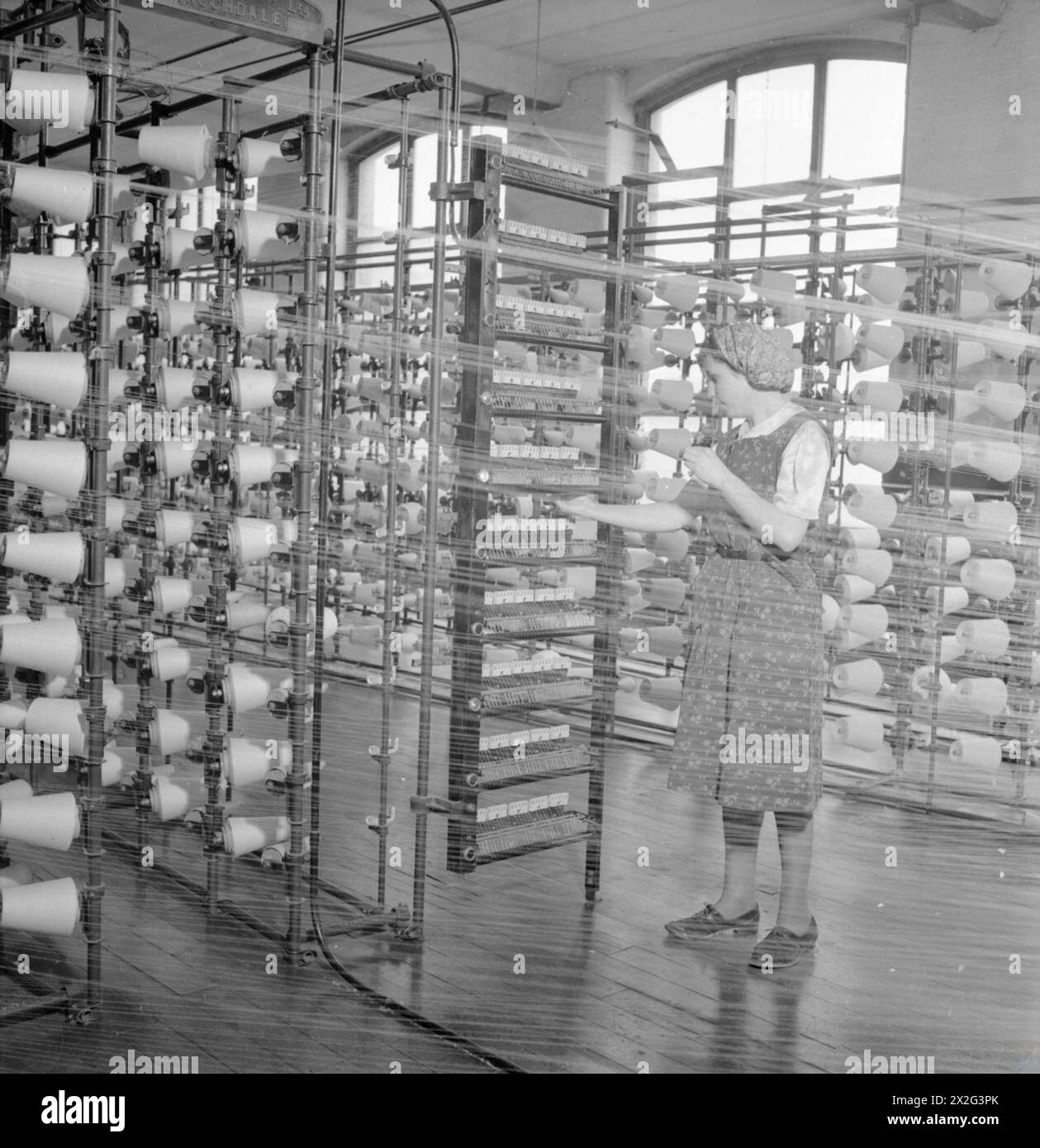 DIE BRITISCHE BAUMWOLLINDUSTRIE: ALLTAG IN Einer BRITISCHEN BAUMWOLLFABRIK, LANCASHIRE, ENGLAND, Großbritannien, 1945 – Hilda Mackenzie arbeitet als Ballwarper in einer Baumwollfabrik irgendwo in Lancashire. Ihre Aufgabe ist es, die feinen Fäden auf einen kreisförmigen Holzbalken aufzuwickeln. Dieses Foto wurde durch das feine Fadengewebe aufgenommen. Reihen und Reihen von Fadenrollen sind deutlich zu sehen Stockfoto