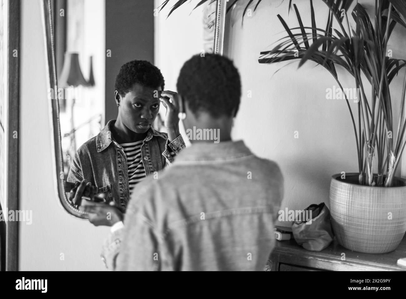 Junge kurzhaarige afrikanische junge Frau schminkt sich vor einem Spiegel und trägt in der Zwischensaison lässige Jeansjacke und Hemd Stockfoto