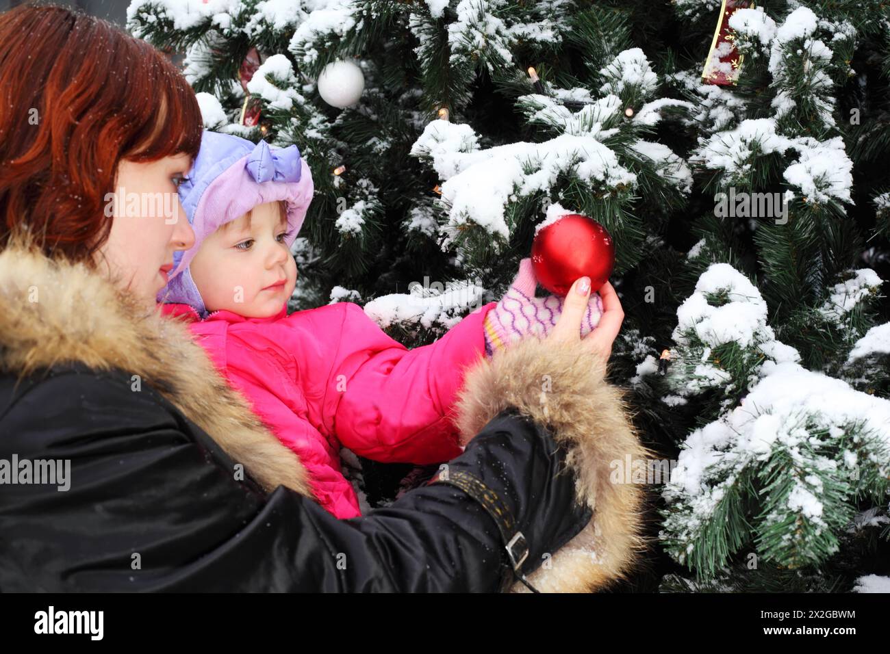 Junge Frau mit kleiner Tochter steht neben einem grünen Baum mit Schnee und rotem Ball Stockfoto